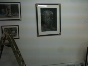 Portrait of face and shoulders, in charcoal. Fragments of other pictures can be seen, with a stepladder in the left foreground