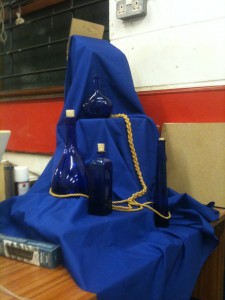 Blue cloth covers platforms of differing levels.  Blue bottles and a gold cord are arranged on it.