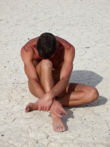 A man, sitting head down, in a close almost foetal position, one knee out towards the ground.
